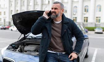an elderly man sits on the hood of a broken car and calls technical assistance on a mobile phone, male insurance agent outdoors photo