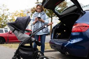 a young mother, using an application on her mobile phone, is trying to figure out how to load and fold a baby stroller into the trunk of a car photo