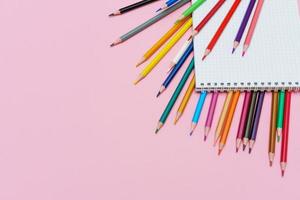 Colored pencils for drawing and a notebook in a cage on a pink background. Back photo