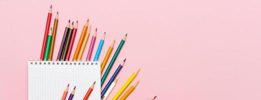 Colored pencils for drawing and a notebook in a cage on a pink background. Back photo