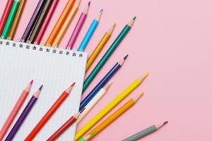 Colored pencils for drawing and a notebook in a cage on a pink background. Back photo