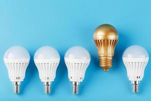 One gold led light bulb is higher and stands out from a row of white lamps on a blue background. photo