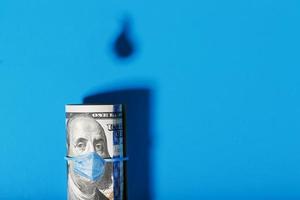 A roll of American bills with a virus mask, with the shadow of oil barrels and a drop of oil on a blue background. photo