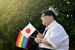 Japanese boy holds rainbow flag and Japan national flag and wears crown and violet neck tie and sitting in front of the tree, soft and selective focus, concept for lgbtq celebration in pride month. photo