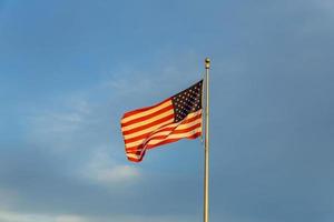 bandera estadounidense en el asta de la bandera ondeando en el viento contra las nubes, el cielo azul y la luna foto