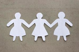 Figures of three girls in dresses holding hands, cut out of white paper. In the center of the photo on a beige background