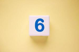 Close-up photo of a white plastic cube with a blue number 6 on a yellow background. Object in the center of the photo