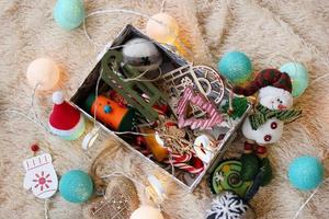 Box with colorful wooden and felt Christmas decorations, caramel sticks and Christmas lights on a soft beige blanket, top view. photo