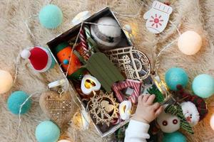 Box with colorful wooden and felt Christmas decorations and Christmas lights with hand of little child on a soft beige blanket, top view. photo