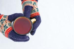 manos de tejer guantes coloridos sostienen una taza de metal con té caliente en el parque de invierno. foto