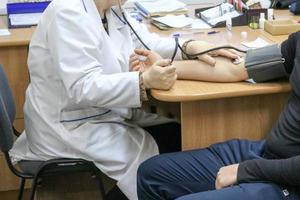 A doctor, a medical worker in a white coat, measures the pressure on the arm of a man sitting on a chair in a medical institution with a tanometer photo