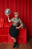 Beautiful stylish mature woman in sunglasses with disco ball and gift box on gift boxes background. Party, celebration, shopping, consumerism concept photo