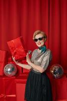 Beautiful stylish mature woman in sunglasses with disco ball and gift box on gift boxes background. Party, celebration, shopping, consumerism concept photo