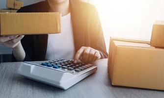 joven y atractiva mujer asiática propietaria de un negocio de inicio mira el trabajo de la cámara feliz con la caja en casa prepara la entrega de paquetes en la cadena de suministro de pymes, adquisiciones, omnicanal o concepto de cuarentena de coronavirus. foto