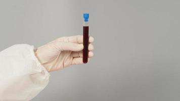 The hand is holding a blood test tube on grey background. Hand wears a PPE suit and a white medical glove. photo