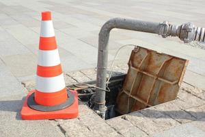 Open manhole, construction traffic warning cone and metal pipe on sidewalk. Concept of repair of supply system, underground utilities, cable laying, water accident. Troubleshooting cleaning sewage. photo