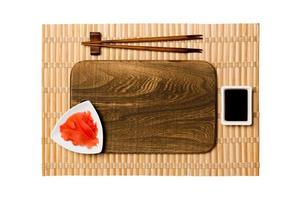 Empty rectangular brown wooden plate with chopsticks for sushi, ginger and soy sauce on yellow bamboo mat background. Top view with copy space for you design photo