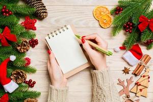 vista superior de la mano femenina escribiendo en un cuaderno sobre fondo de Navidad de madera. abeto y decoraciones festivas. lista de deseos concepto de año nuevo foto