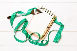 Fitness concept with empty space for your design on white wooden background. Top view of notepad and fork with green measure tape photo