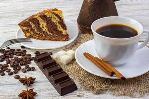 Delicious chocolate cake on a plate with a cup of coffee . Coffee beans, a piece of chocolate, anice, sugar and cinnamon sticks on wooden table