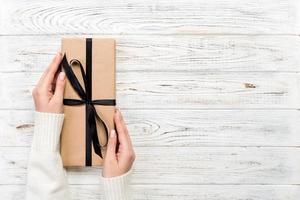 Woman hands give wrapped valentine or other holiday handmade present in paper with black ribbon. Present box, decoration of gift on white wooden table, top view with copy space photo