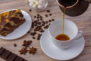 A cup of coffee and a tasty cake on a saucer. Chocolate bar, coffee beans, a bowl with sugar cubes, and anise on bright wooden background. Top view photo