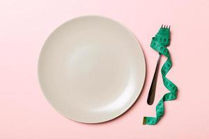 Strict diet concept with empty space fro your design. Top view of plate with fork in measuring tape on pink background photo