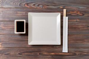 Empty white square plate with chopsticks for sushi and soy sauce on wooden background. Top view with copy space photo
