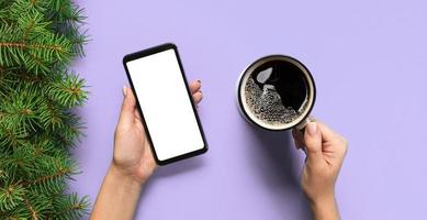 Female hands holding black mobile phone with blank white screen and mug of coffee. Mockup banner image with copy space. Top view on purple background, flat lay photo