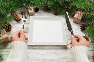 vista en perspectiva la niña sostiene un tenedor y un cuchillo en la mano y está lista para comer. plato cuadrado blanco vacío sobre fondo de Navidad de madera. concepto de plato de cena navideña con decoración de año nuevo foto