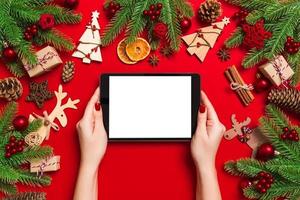 Top view of woman holding tablet in her hands on red background made of Christmas decorations. New Year holiday concept. Mockup photo