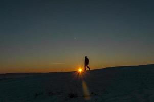 silueta de un hombre al atardecer caminando en invierno, poniendo el sol detrás de la montaña foto