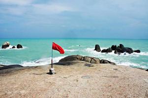 Red flag standing on the rocky coast of the Gulf of Thailand against the backdrop of the blue sea. Hazard warning during storm photo