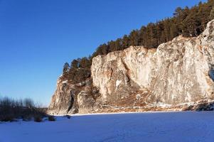 Winter mountain landscape with cliffs on a frozen and snow-covered river. Rocks with pine trees, scenic nature photo