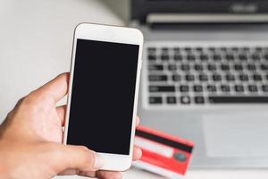 mano izquierda usando un teléfono inteligente en un fondo negro borroso con una computadora portátil y una tarjeta de crédito foto