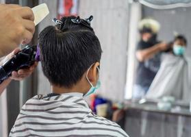 un adolescente con una máscara protectora facial se está cortando el pelo en una barbería. cortes de pelo alargados de moda para niños. salón de belleza en cuarentena coronavirus covid-1 foto
