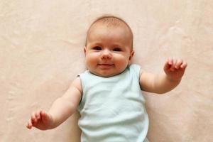 el retrato de un bebé sonriente de tres meses con traje azul está acostado sobre una manta beige y mirando la cámara. foto