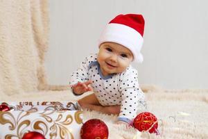 Adorable smiling little girl in Santa Claus red hat is playing with red shiny Christmas decorations from a box on a beige plaid with Christmas lights. photo