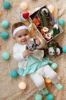 Beautiful smiling little girl in blue and white dress is playing with felt ball on a beige plaid with Christmas decorations and lights, top view. photo