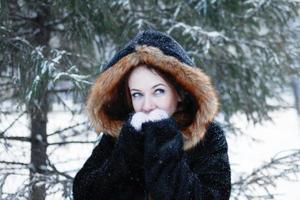 Portrait of young beautiful woman with blue eyes in black faux fur coat with red hood on background of winter snowy park. photo