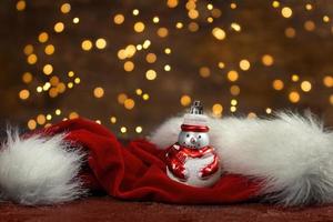 Snowman and red santa claus hat with new year lights in the back. Blurred background. Christmas card photo