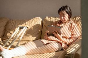 Girl with a cast on their leg, sitting on the couch and drawing on a graphics tablet. Bandaged leg cast and toes after a running injury accident.Teen girl in a plaster cast. photo