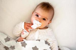 retrato sincero de una linda niña caucásica acostada en una cama blanca con chupete y juguete de liebre. foto