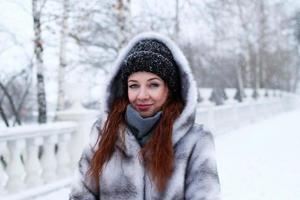 joven mujer hermosa con pelo rojo y ojos azules en abrigo de piel sintética gris con capucha en el fondo del parque nevado de invierno. foto