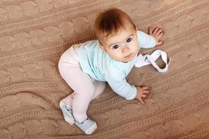 hermosa niña con ojos azules y cabello rubio está sentada en una manta de punto beige y mirando a la cámara. foto