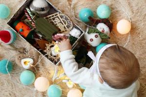 Little girl in white dress is trying to take Christmas decorations from a box, top view. Waiting for New Year and Christmas holidays. photo