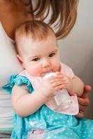 Cute little Caucasian baby girl in blue dress drinking water from bottle on the mother's knees. Child learns to hold a bottle. photo