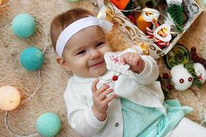 hermosa niña sonriente con vestido azul y blanco está jugando con un juguete de madera en una manta beige con decoraciones y luces navideñas, vista superior. foto