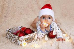 Cute little girl in Santa Claus red hat is playing with Christmas balls and Christmas lights on a beige plaid near to box with red and white shiny Christmas decorations. photo