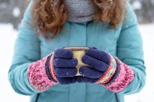 las manos de las mujeres en tejer guantes coloridos sostienen una taza de metal con té caliente en el parque de invierno. foto